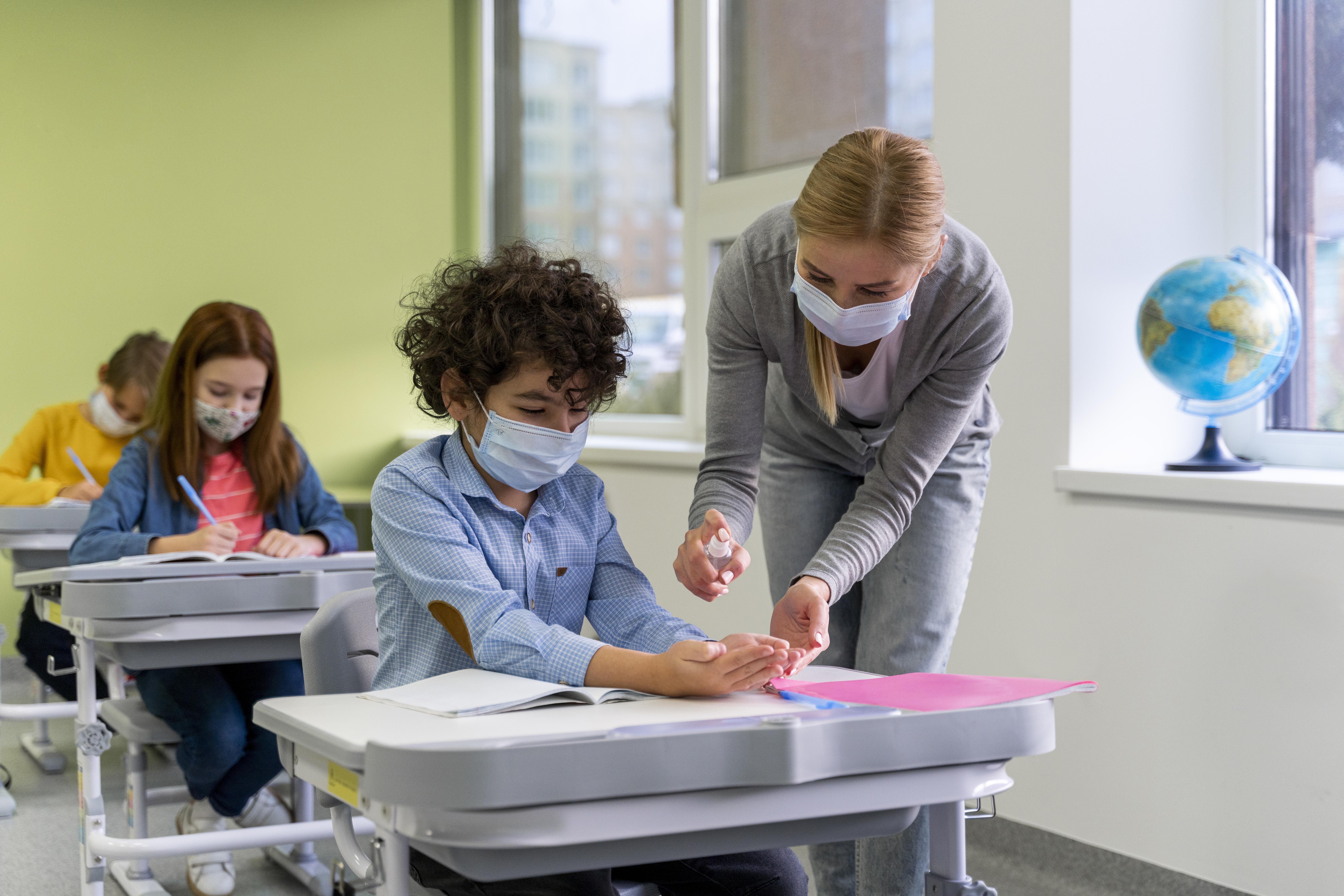 female-teacher-with-medical-mask-giving-hand-sanitizer-children-classroom