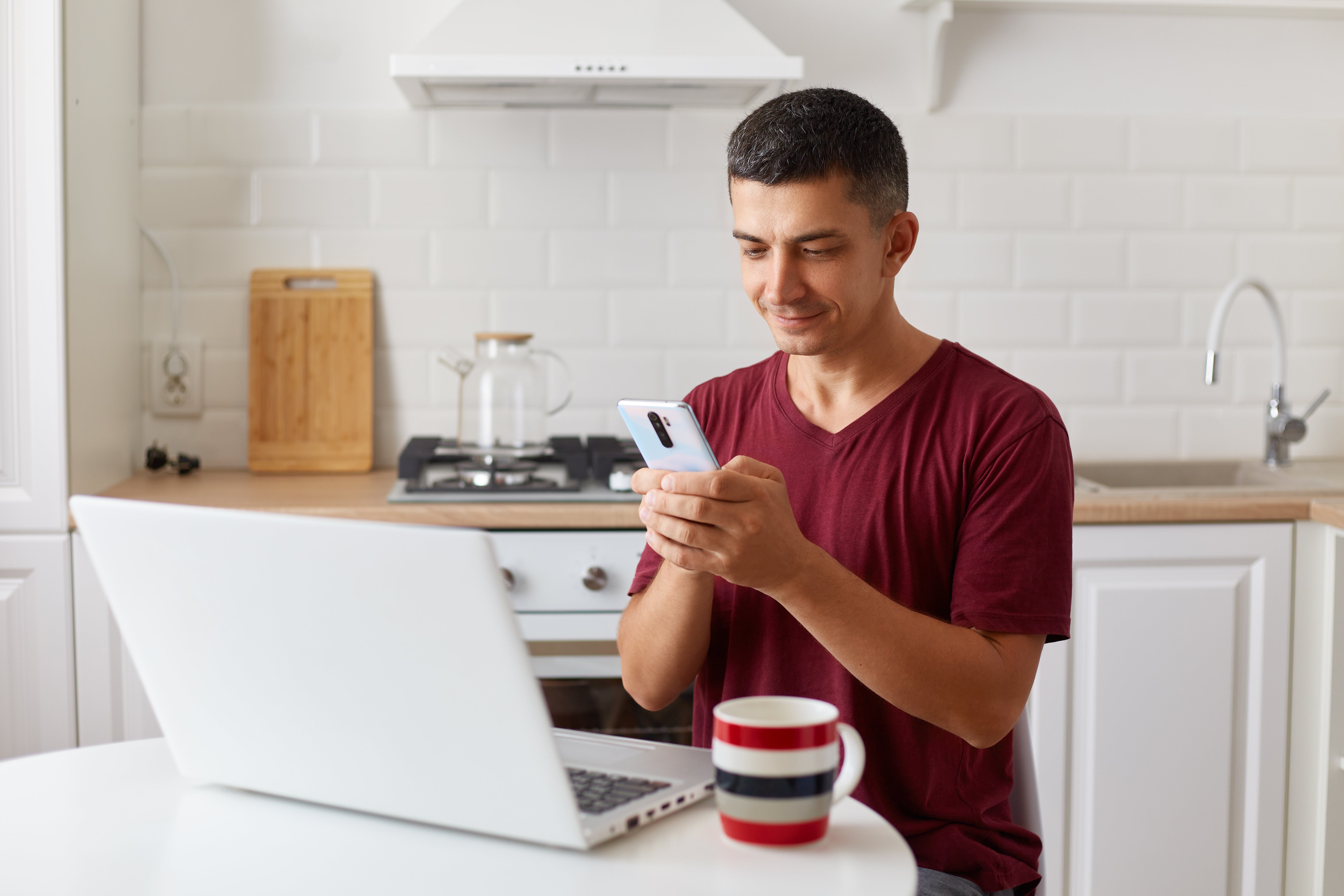 positive-handsome-man-working-online-home-laptop-using-smartphone-checking-e-mails-while-having-break-fro-freelance-work-looking-device-screen-typing-message