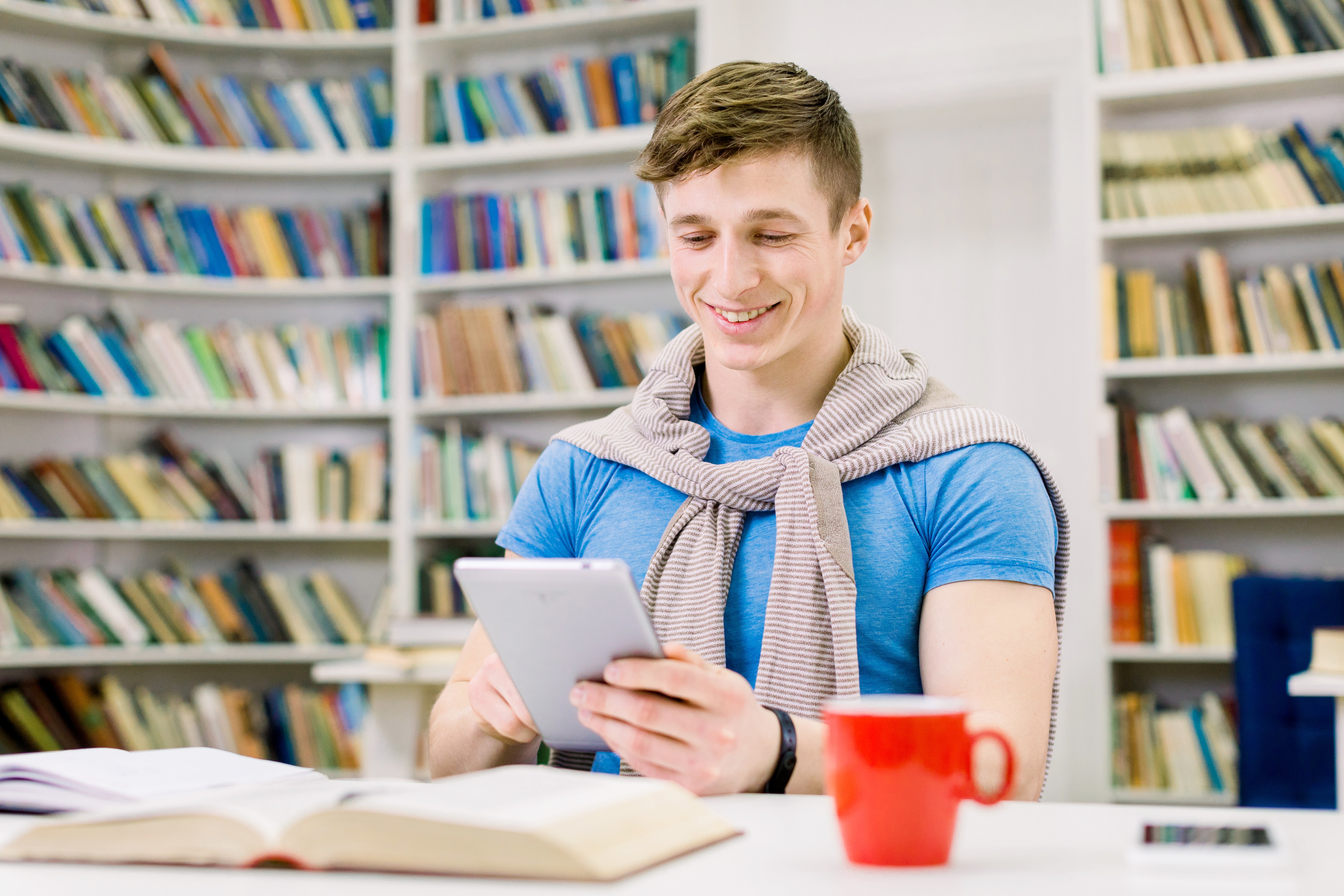 smiling-satisfied-handsome-young-male-student-sitting-library-searching-information-i-pad-during-preparation-test