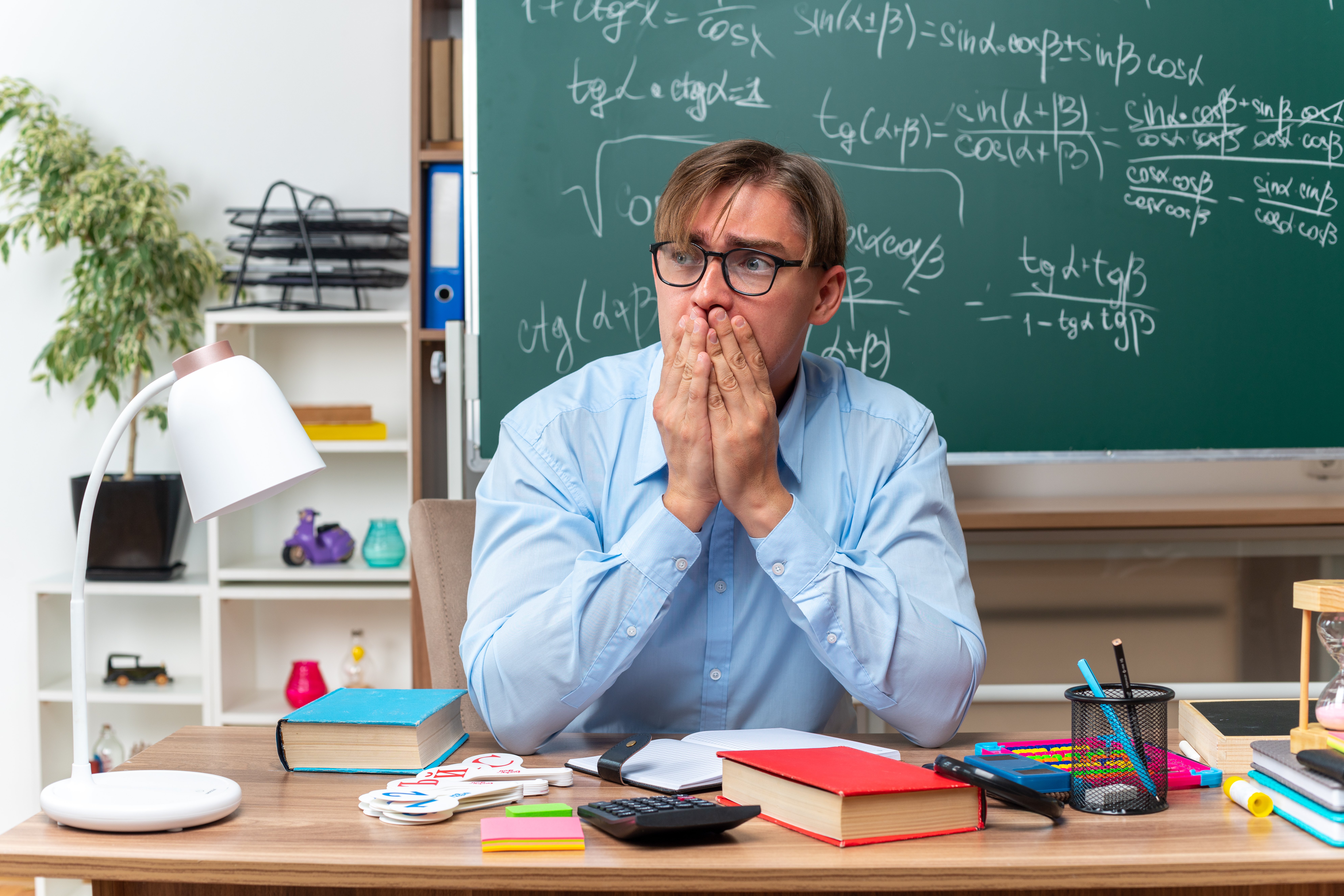 young-male-teacher-wearing-glasses-looking-aside-stressed-nervous-covering-mouth-with-hands-sitting-school-desk-with-books-notes-front-blackboard-classroom